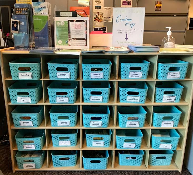 The Well's Condom Co-Op station is a wooden shelf of individually-labelled blue bins stocked with safer sex supplies